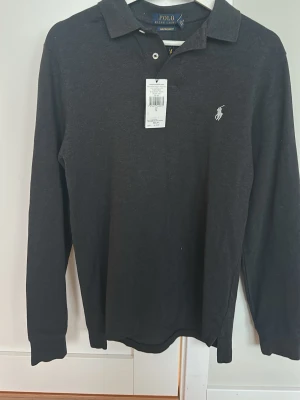 Mörkgrå långärmad pikétröja Polo Ralph Lauren - Klassisk mörkgrå långärmad pikétröja från Polo Ralph Lauren med vit broderad logga på bröstet. Tröjan har krage, knappar vid halsen och är tillverkad i mjuk bomull. Perfekt för en stilren och avslappnad look. Tröjan är köpt i ralph lauren butik i Spanien och har ej använts sedan eftersom köpt som en present men passade ej 