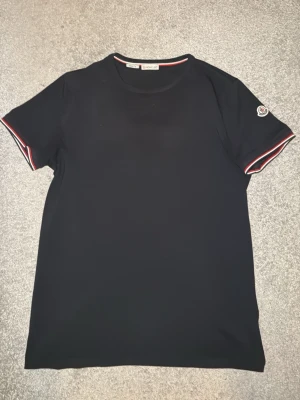 Mörkblå moncler polo t shirt  - Svart t-shirt från Moncler i slim fit-modell. T-shirten har korta ärmar och detaljer med rödvita ränder vid ärmslut samt Moncler-logga på ena ärmen. Tillverkad i mjuk bomull för en skön känsla.äkta moncler polo t shirt köpt från nk har kvitto 