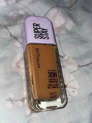 Maybelline Super Stay Foundation 337 - Maybelline Super Stay 30H Lumi-Matte Foundation i nyansen 337. Kommer i en rektangulär glasflaska med lila plastlock och svart text. Ger en matt finish och lång hållbarhet. Perfekt för dig som vill ha en täckande bas med naturlig look.
