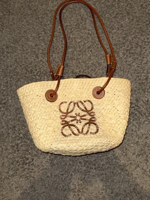 Stråväska med bruna läderdetaljer - Snygg stråväska från Loewe med bruna läderhandtag och dekorativt brunt lädermönster på framsidan. Väskan har en klassisk korgform och är tillverkad i flätad naturfärgad strå. Perfekt för dig som gillar en trendig och avslappnad stil.