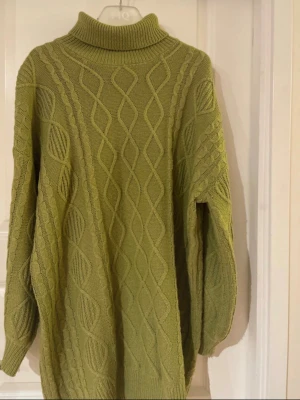 Grön polotröja med kabelstickning - Stickad polotröja i olivgrönt med snygg kabelstickad design och bred polokrage. Tröjan har lång ärm och avslappnad passform, perfekt för lager på lager. Materialet ser ut att vara mjukt och värmande, troligen bomull eller ullblandning.