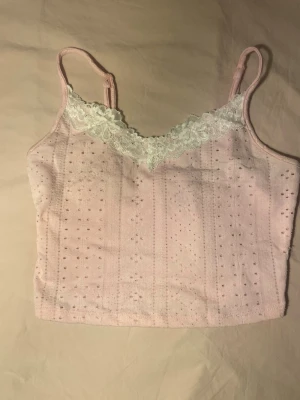 Ljusrosa spetsdetaljerad linnetopp - Söt ljusrosa linnetopp med smala axelband och v-ringning dekorerad med vit spets. Toppen har ett hålmönster i tyget och är croppad, perfekt för en trendig look. Materialet känns mjukt och lätt.