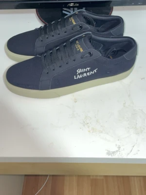 Svarta Saint Laurent sneakers - Svarta sneakers från Saint Laurent med vit sula och klassisk låg profil. Skorna har textlogga på sidan och gulddetaljer på plösen. Tillverkade i canvas med matchande svarta skosnören. Perfekta för en clean och stilren look. Kvitto finns för äktahets bevis