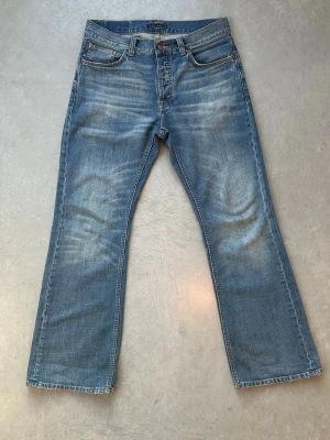 Flare Glenn Nudie Jeans blåa - Säljer ett par klassiska blå bootcut jeans från Nudie Jeans med snygg slitning och kontrastsömmar. Jeansen har fem fickor, knappgylf och är tillverkade i mjuk denim. Perfekta för dig som gillar en avslappnad och tidlös stil. Modelnamnet är flare Glenn. Storlek 31 W 32 L. Väldigt nöja med de men är för stora i midjan för mig. 