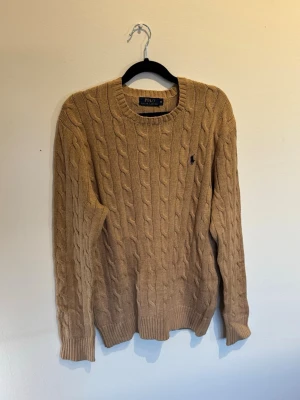 Beige kabelstickad tröja Polo Ralph Lauren - Ralph Lauren Stickad. Mycket fint skick, Storlek M Kabelstickad tröja i beige från Polo Ralph Lauren med rund halsringning och mörkblå broderad logga på bröstet. Tröjan har ribbade muddar vid ärmslut och nederkant. Perfekt för dig som gillar klassisk och stilren look.