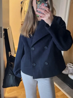 Mörkblå dubbelknäppt rock från H&M - Kort, mörkblå rock från H&M med dubbelknäppning och bred krage. Klassisk boxig passform och raka ärmar. Tillverkad i ett tjockare material som ger en stilren look. Perfekt för kyligare dagar och enkel att matcha med olika outfits.