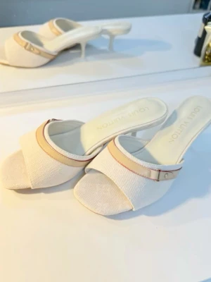 Louis Vuitton vita sandaler - Snygga vita sandaler från Louis Vuitton med låg klack och öppen tå. Skorna har en beige detalj med guldfärgad knapp och diskret rosa söm. Materialet är skinn med textilstruktur och logotypen är präglad på innersulan. Perfekt för dig som vill ha en lyxig och stilren look❤️INTE ÄKTA
