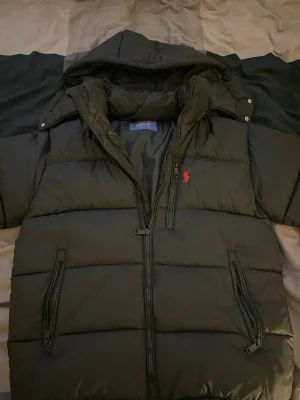 Svart dunjacka från Polo Ralph Lauren - Snygg svart dunjacka från Polo Ralph Lauren med huva och två dragkedjeförsedda fickor framtill. Jackan har det klassiska röda Polo-logotypbroderiet på bröstet och quiltad design. Perfekt för kalla vinterdagar och håller dig varm med stil.