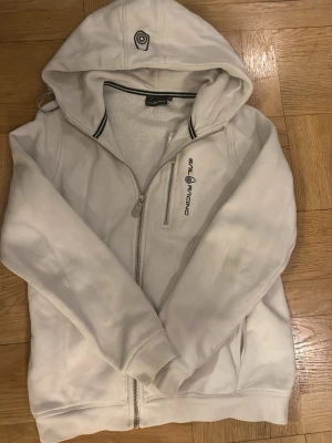 Vit hoodie från Sail Racing - Vit hoodie från Sail Racing med dragkedja och broderad logga på bröstet. Mjuk insida i fleece och praktisk huva. Snygga detaljer med svarta ränder på kragen och fickor på sidorna. Perfekt för chill dagar eller när du vill ha en clean look. Passar om man är 160cm.