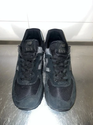 Svarta New Balance 574 sneakers - Svarta New Balance 574 sneakers med klassisk siluett, mesh och mockadetaljer samt reflekterande N-logga på sidorna. Skorna har grov yttersula för bra grepp och svart snörning. Perfekta för dig som gillar stilrena och sportiga sneakers. Sköna och feta skor!!! 