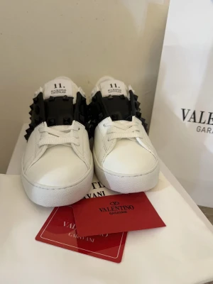 Valentino Garavani vita sneakers - Säljer ett par vita sneakers från Valentino Garavani med svart band och svarta nitar på sidorna. Skorna har en klassisk låg siluett, vita snören och är tillverkade i slätt läder. Perfekta för dig som vill ha en edgy och exklusiv look.