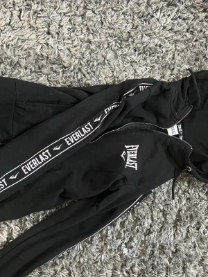Svart Everlast hoodie med dragkedja - Säljer en svart hoodie från Everlast med dragkedja framtill och vita logotexter längs ärmarna och bröstet. Hoodien har huva med dragsko och ribbade muddar. Perfekt för en sportig och avslappnad stil.