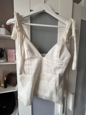 Vit linnetopp med knyt från Zara - Snygg vit linnetopp från Zara med breda axelband som knyts upptill. Toppen har v-ringning och figurnära passform. Materialet känns mjukt och lätt, vilket gör den skön att bära under varma dagar.