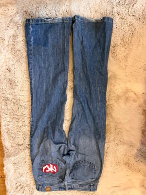Blå bootcut jeans från Replay - Säljer ett par klassiska blå bootcut jeans från Replay med fem fickor och låg midja. Jeansen har en snygg tvätt och NY äpple logga i rött på bakfickan. Perfekta för dig som gillar retrostil och vill ha ett par jeans med lite vidare ben. Köpte dem från tinlessel. Mått: midjemått:40cm midjehöjd:18 cm innebenslängd:79cm ytterbenslängd:99cm benöppning:25cm 