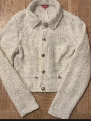 Vit kavajjacka i bouclé med knappar - Stilren vit kavajjacka i bouclé med markerad krage och dekorativa silverfärgade knappar framtill. Jackan har lång ärm, croppad passform och två fickdetaljer. Perfekt för en chic och trendig look.