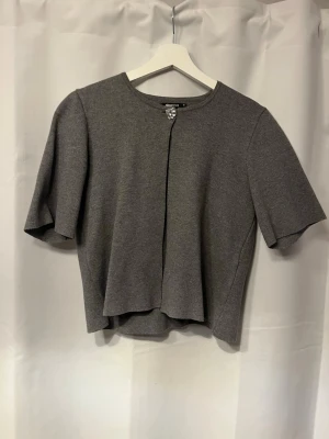 Grå kortärmad kofta från Gina Tricot - Stilren grå kofta från Gina Tricot med korta ärmar och rund halsringning. Koftan har en dekorativ knapp framtill vid halsen och är tillverkad i ett mjukt stickat material. Perfekt att slänga över en topp för en enkel och clean look. Köptes för 400 kr, använt 5-10 ggr💕