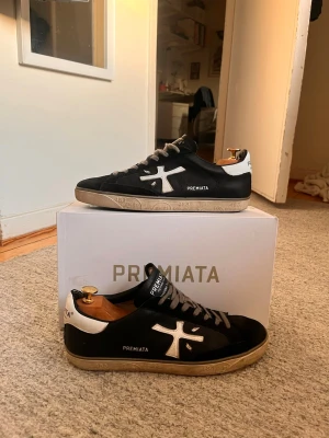 Svarta Premiatas - Snygga svarta sneakers från Premiata med vita detaljer och logga på sidan. Skorna har en låg silhuett, vita skosnören och beige sula med tryck. Ovandelen är i mocka och läder, och hälen har en vit kontrast. Perfekta för en trendig streetstyle-look. Fint skick har ändast används i 2 månader men har tyvärr gått upp i insidan av skon. Går att laga ganska lätt. Annars är dom i superbra skick. Köptes för 3200 allt og medföljes förutom kvittot eftersom det inte finns kvar!
