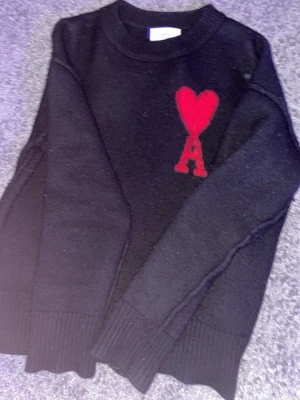 Svart stickad tröja från Ami Paris - Säljer en svart stickad tröja från Ami Paris med deras ikoniska röda hjärta och bokstaven A broderat på bröstet. Tröjan har rund halsringning, långa ärmar och ribbade muddar. Perfekt för dig som gillar stilrena plagg med en twist.