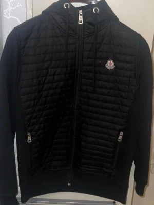 Svart hoodie från Moncler med dragkedja - Snygg svart hoodie från Moncler med quiltad front och rygg, dragkedja hela vägen och två fickor med dragkedja. Klassisk Moncler-logga på bröstet och justerbar huva med snören. Perfekt för dig som vill ha en stilren och exklusiv look.