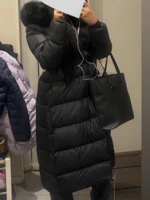 Lång svart dunjacka med pälskrage - Säljer en lång, svart dunjacka med stor fluffig pälskrage på huvan. Jackan har en quiltad design med breda paneler och dragkedja framtill. Perfekt för kalla vinterdagar och riktigt snygg till vardagslooken.