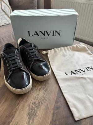 Svarta Lanvin sneakers i mocka och lack - Snygga svarta sneakers från Lanvin med ovandel i mocka och tåparti i blankt lack. Klassisk låg modell med vita sulor och svarta skosnören. Perfekta för dig som vill ha en stilren och exklusiv sneaker med lyxig känsla.