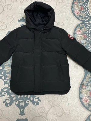 Svart dunjacka från Canada Goose - Säljer en svart dunjacka från Canada Goose med huva och klassisk logotyp på ärmen. Jackan har två sidofickor och en stilren, quiltad design som håller dig varm under kalla dagar. Perfekt för dig som vill ha både funktion och stil. Size is S and condition is perfect. Used couple times. 