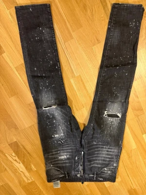 Svarta dsquared2 jeans med slitningar - Coola svarta jeans med slitningar och paint splatter-effekt. De har raka ben, trasiga detaljer på knäna och klassiska fem fickor. Materialet är denim och jeansen har en modern, trendig look som passar perfekt för dig som gillar streetstyle. Väldigt fina köpa i köpemhamn tags finns och allt