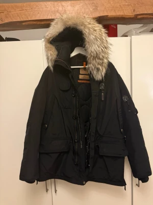 Svart dunjacka med pälskrage Parajumpers - Säljer en svart dunjacka från Parajumpers med stor huva och avtagbar pälskant. Jackan har flera fickor, dragkedja och knappar framtill samt detaljer som orange hängare och patch på ärmen. Perfekt för kalla vinterdagar. Den är i väldigt bra skick, nyss lagt in den i kemtvätt också, köpt en ny vinterjacka så vill bli av med denna. Det är svårt o hitta så här fin päls på jackorna.