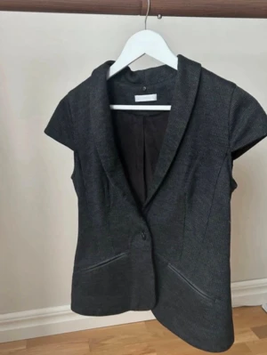 Mörkgrå väst från Promod - Snygg mörkgrå väst från Promod med markerad krage och en knapp framtill. Västen har två fickor och är tillverkad i ett lätt strukturerat material. Perfekt för att addera en stilren touch till din outfit.