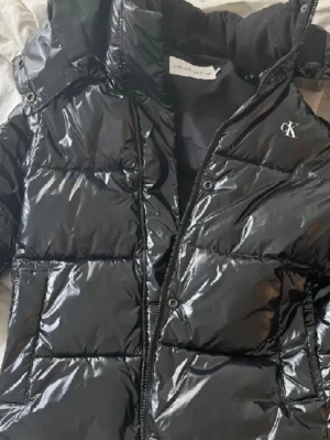 Svart glansig dunjacka Calvin Klein - Svart glansig dunjacka från Calvin Klein med huva och dragkedja. Jackan har CK-logga på bröstet och Calvin Klein Jeans-tryck på ärmen. Puffad design med stora paneler och praktiska fickor framtill. Perfekt för kalla dagar.