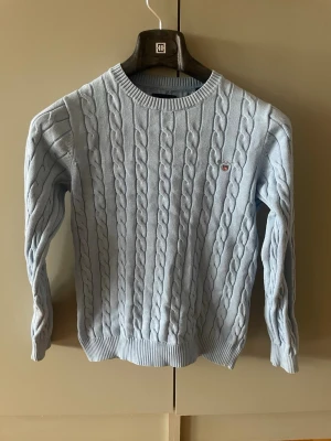 Ljusblå kabelstickad tröja Gant - Snygg ljusblå kabelstickad tröja från Gant i premium bomull. Tröjan har rund halsringning, ribbade muddar och en liten Gant-logga broderad på bröstet. Perfekt för dig som gillar klassisk och stilren look.