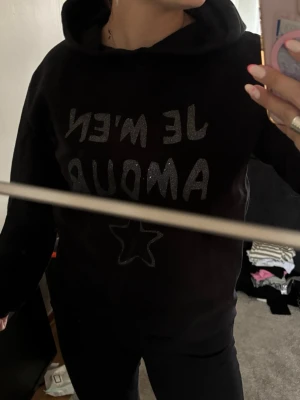 Svart hoodie med text och stjärna - Säljer en svart hoodie med huva och glittrig texten 'JE M'EN AMOUR' på bröstet samt en stjärna under texten. Hoodien har en avslappnad passform och är perfekt för en chill och trendig look. Materialet känns mjukt och bekvämt. Barnstorlek men skulle säga xs 