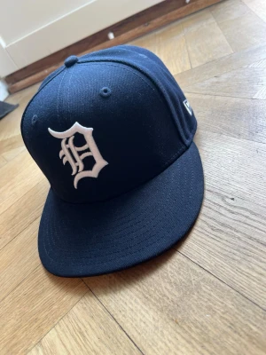 Mörkblå New Era 59FIFTY keps - Säljer en mörkblå New Era 59FIFTY keps med Detroit Tigers-logga i vitt framtill. Klassisk flat brim och broderade ventilationshål. Tillverkad i ett slitstarkt material och officiell MLB On-Field modell. Perfekt för dig som gillar sportig streetstyle.