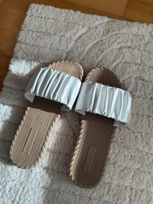 Beige sandaler med plisserad rem - Säljer ett par trendiga sandaler i beige med en bred, vit plisserad rem över foten.