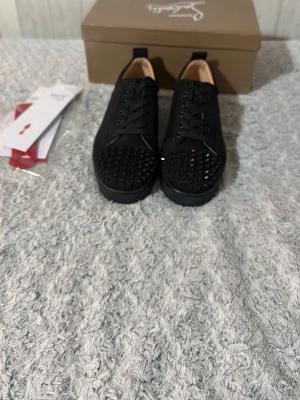 Svarta sneakers med nitar från Louboutin - Säljer ett par svarta sneakers från Christian Louboutin med coola svarta nitar på tån. Skorna har klassisk låg profil, svarta skosnören och en stilren design i läder och textil. Perfekta för dig som vill sticka ut med en edgy look.
