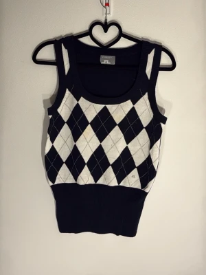 Argyle stickad väst från J.Lindeberg - Snygg stickad väst från J.Lindeberg i svart och vitt argylemönster. Modellen är ärmlös med breda axelband och ribbad nederkant. Perfekt att styla över en skjorta eller t-shirt för en trendig look.