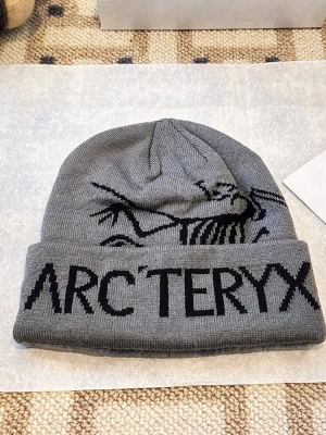 Grå mössa från Arc'teryx - Snygg grå mössa från Arc'teryx med svart logga och fossilmönster på framsidan. Mössan är ribbstickad och har en uppvikt kant med tydlig branding. Perfekt för dig som gillar outdoor och streetwear vibes.