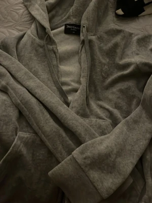 Grå velour hoodie från Juicy Couture - Mjuk och skön grå hoodie från Juicy Couture i velourmaterial. Klassisk huva, dragkedja framtill och fickor på sidorna. Diskret Juicy Couture-logga broderad på bröstet. Perfekt för en avslappnad och trendig look.