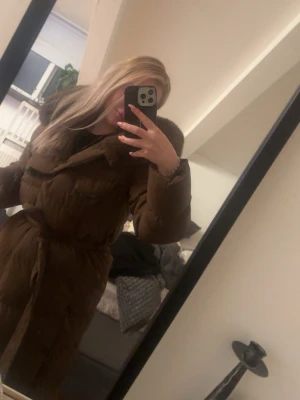 Brun lång dunjacka med äkta kanin päls - Säljer en brun lång dunjacka med stor huva och äkta kanin päls. Jackan har knytband i midjan för snygg passform och stora fickor framtill. Perfekt för kalla vinterdagar och riktigt mysig att bära utomhus.