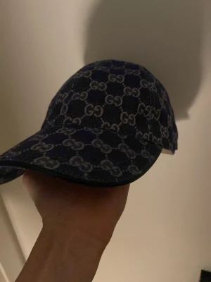 Mörkblå Gucci keps med GG-mönster - Snygg mörkblå keps från Gucci med det klassiska GG-monogrammet över hela kepsen. Kepsen har böjd skärm och justerbart spänne bak. Materialet ser ut att vara denim eller canvas, perfekt för en trendig streetstyle-look.
