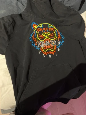 Svart Kenzo hoodie med tigertryck - Svart hoodie från Kenzo med ikoniskt broderat tigerhuvud i neonfärger och texten 'KENZO PARIS' på bröstet. Klassisk känguruficka framtill och huva. Perfekt för dig som vill sticka ut med en streetwear-look.