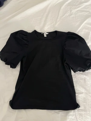 Svart topp med puffärmar - Snygg svart topp med puffiga kortärmar och rund halsringning. Toppen har en stilren design och är gjord i mjukt bomullstyg med ärmar i ett glansigare material. Perfekt för dig som vill ha en trendig och bekväm look.