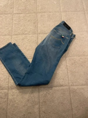 Blå slim fit jeans från Jacob Cohen - Säljer ett par blå slim fit jeans från Jacob Cohen med klassisk femficksmodell och snygga slitningar framtill. Jeansen har en liten italiensk flaggdetalj på bakfickan och är tillverkade i mjukt denimtyg. Perfekta för en avslappnad och trendig look.