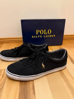 Svarta Polo Ralph Lauren sneakers - Snygga svarta sneakers från Polo Ralph Lauren med klassisk låg profil och vit sula. Skorna har gul broderad logga på sidan och är tillverkade i canvasmaterial. Perfekta för en clean och stilren look.