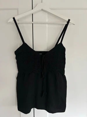 Svart virkad linnetopp med knyt - Snygg svart linnetopp med virkade detaljer upptill och smala axelband. Toppen har knyt framtill och en lätt vågig kant under bysten. Perfekt till sommaren och enkel att styla med jeans eller kjol.