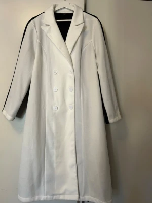 Vit lång dubbelknäppt kappa - Snygg vit kappa i lång modell med dubbelknäppning och fyra vita knappar framtill. Klassisk krage och raka ärmar. Materialet ser ut att vara mjukt och ull-liknande, perfekt för en clean och elegant stil.