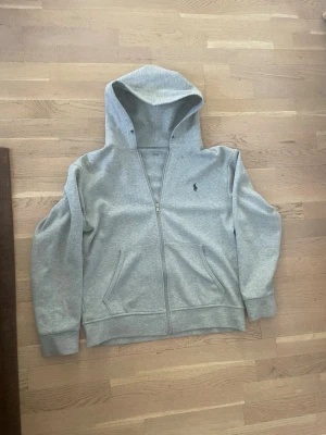 Grå zip hoodie från Polo Ralph Lauren - Snygg grå hoodie med dragkedja från Polo Ralph Lauren. Klassisk design med huva, två fickor fram och den ikoniska svarta polospelaren broderad på bröstet. Tillverkad i mjukt bomullsmaterial som är perfekt för chill dagar.