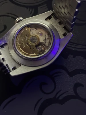 Seiko automatisk klocka i silver - Snygg automatisk klocka från Seiko i helsilver med klassisk länkarmband och räfflad bezel. Urtavlan är mörk med Arabiska nummer och tydliga index. Baksidan har genomskinligt fönster som visar urverket. Perfekt för dig som gillar stilrena accessoarer.40mm ur. finns box länkar och verktyg för att öppna den vid behov 