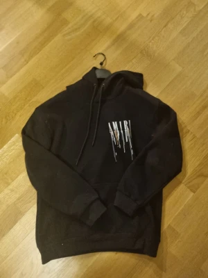 Svart amiri hoodie - Säljer en svart hoodie från Amiri med stor huva och dragsko. På bröstet och ryggen finns ett vitt AMIRI-tryck med färgstänk i orange, blått och vitt som ger en cool streetwear-vibe. Hoodien har magficka och ribbade muddar. Perfekt för dig som gillar snygga detaljer. Storlek S inga defekter. Fraktar fort