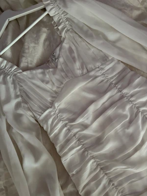 Vit omlottklänning med långa ärmar - Säljer en vit kortklänning i omlottmodell med långa, transparenta ärmar och rynkade detaljer längs hela klänningen. Materialet har en sidenliknande finish som ger ett lyxigt intryck. Perfekt för dig som vill ha en elegant och trendig look.